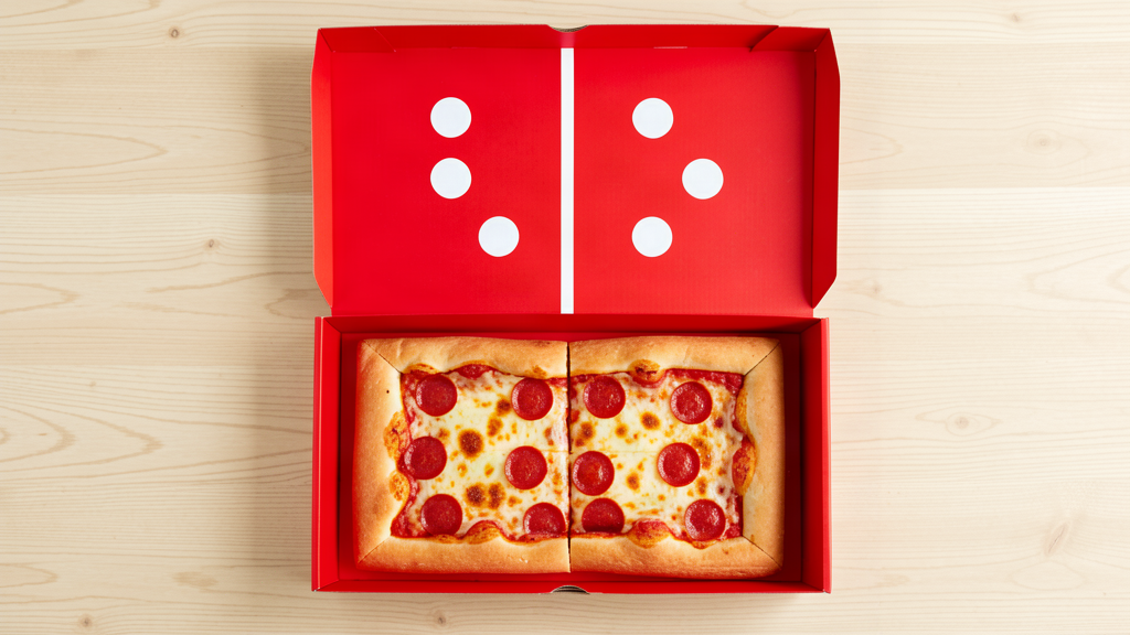 Top-down view of a red domino-style pizza box, open, with a white domino face on the lid, showing a pizza cut into two slices inside